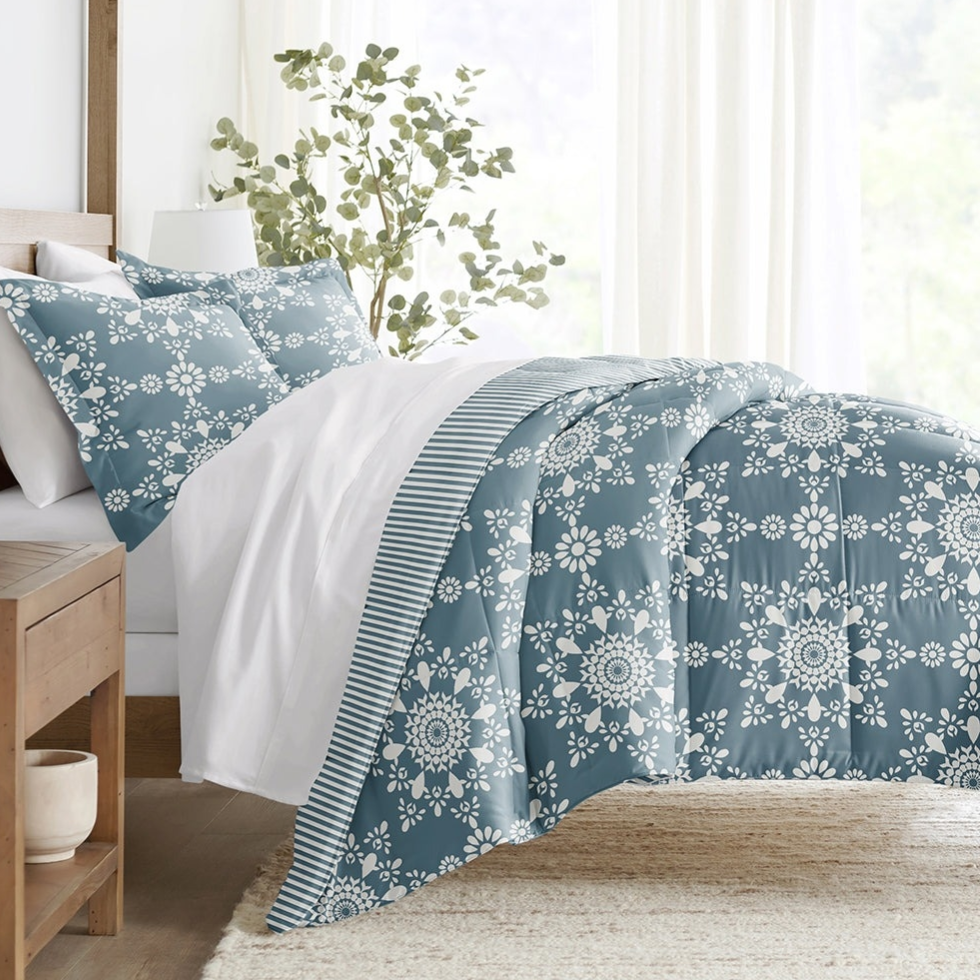 A bed features a blue and white floral-patterned comforter and pillowcases, with a striped accent on the comforter's edge, set against a white bedsheet. A wooden nightstand with a small vase and a leafy plant in the background complete the decor.