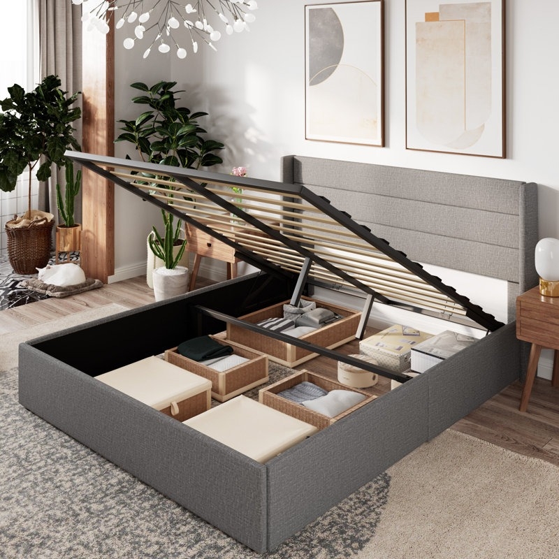 A grey upholstered platform bed with a gas lift mechanism reveals under-bed storage, featuring organized compartments with baskets. The room includes plants, abstract artwork, and a contemporary chandelier.