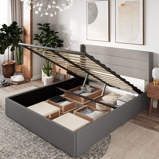 A grey upholstered platform bed with a gas lift mechanism reveals under-bed storage, featuring organized compartments with baskets. The room includes plants, abstract artwork, and a contemporary chandelier.