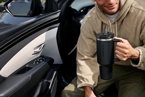 A person is sitting in a car holding a large black Stanley travel mug.