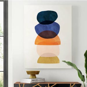 The Mercury Row Mod Pods Print features a series of stacked, colorful semi-circular shapes in blue, orange, brown, and gold hues on a white background. It decorates a modern interior space with a plant, books, and a decorative bowl.