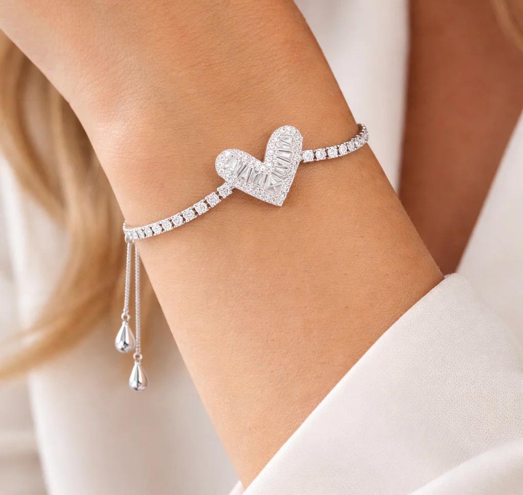 A silver bracelet features a heart-shaped pendant with baguette and round-cut stones, accompanied by an adjustable chain and teardrop-shaped ends.