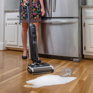 A handheld cordless vacuum with an upright build featuring a cleaning head at its base is depicted in use on a kitchen floor where a liquid spill is present.