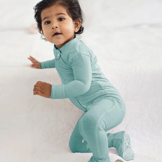 A baby is wearing a teal one-piece romper with foot grips, sitting on a white surface.
