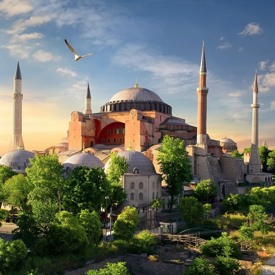 A large, historic building with a central dome and four minarets surrounded by lush greenery, set against a serene sky and featuring a bird in flight.