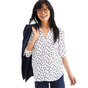 A woman is wearing a white Johnny Collar shirt with navy polka dots, featuring a V-neck and long sleeves, paired with navy pants. She holds a dark blazer draped over her shoulder, adding a professional touch.