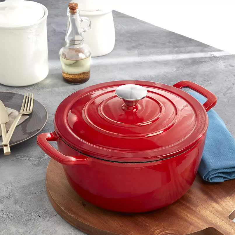 Red enameled cast iron Dutch oven with a silver knob on a wooden cutting board, surrounded by a blue napkin, white ceramic canisters, a bottle of oil, a plate, and utensils.