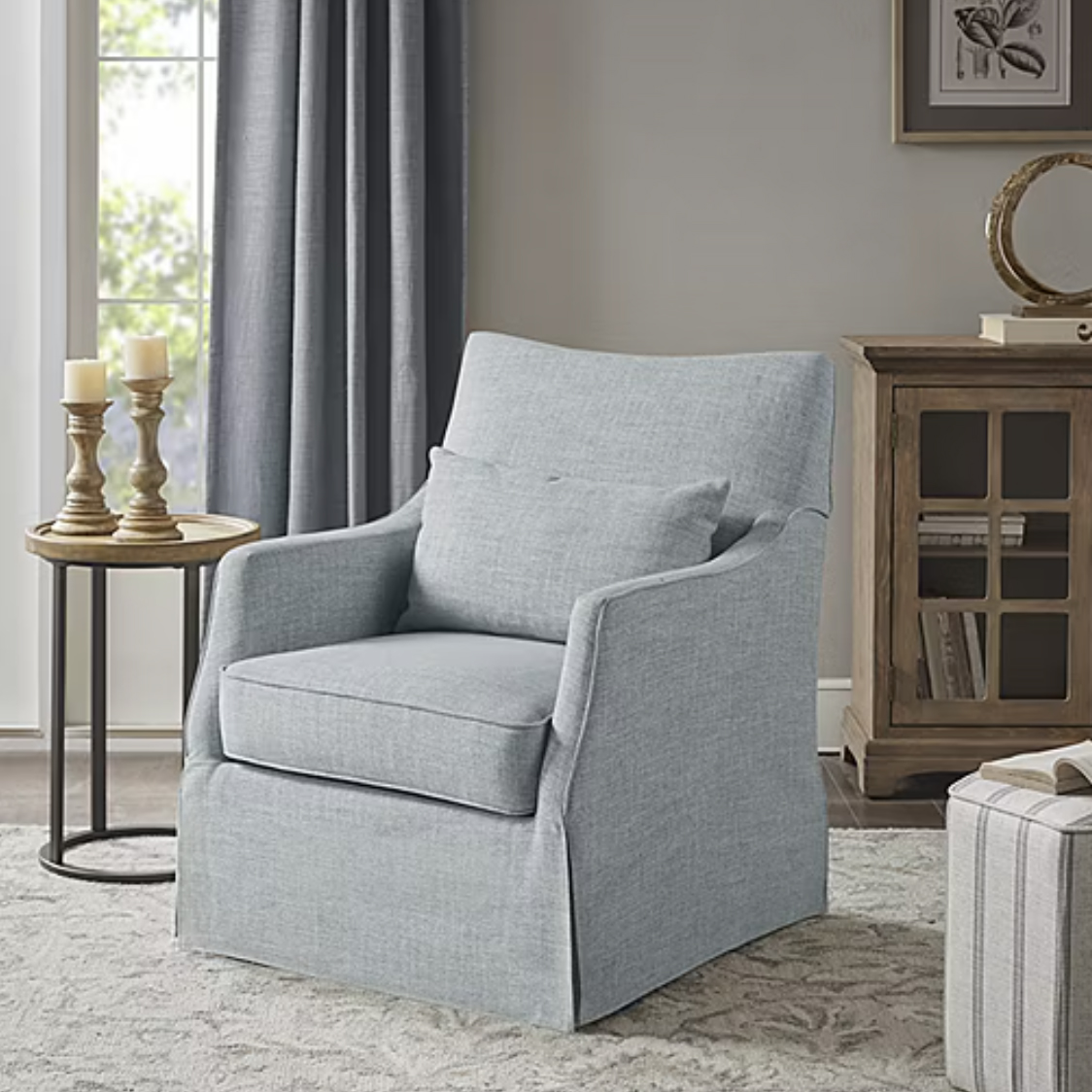 A light gray upholstered armchair with a modern design, featuring a cushioned seat and back, and slim armrests. It sits on a thick rug next to a round side table with candles and is near a wooden cabinet and window with curtains.