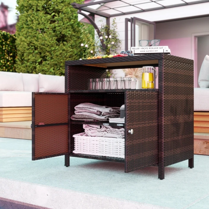 Outdoor storage cabinet with a dark wicker design, featuring open shelves and closed compartments. It holds glasses, a jug, folded towels, and a basket. There's a book and food items on top, with a patio setting in the background.
