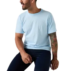 A person wearing a light blue T-shirt and dark pants sits on a white surface, showcasing a colorful tattoo on their left arm.