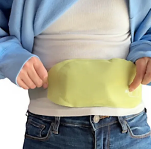 A person wears a portable yellow heating pad around their waist, secured outside a white shirt and under a blue jacket, providing warmth and relief.