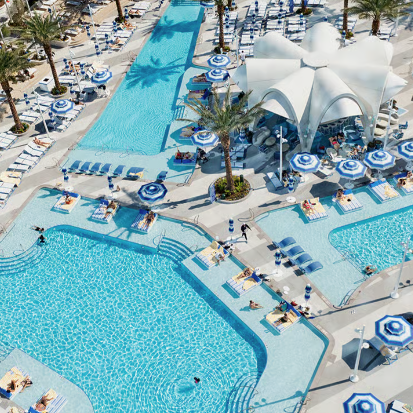 A large, luxurious swimming pool complex features palm trees, blue-striped umbrellas, numerous sun loungers, and a central tent-like canopy structure, creating a resort-style setting.