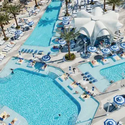 A large, luxurious swimming pool complex features palm trees, blue-striped umbrellas, numerous sun loungers, and a central tent-like canopy structure, creating a resort-style setting.