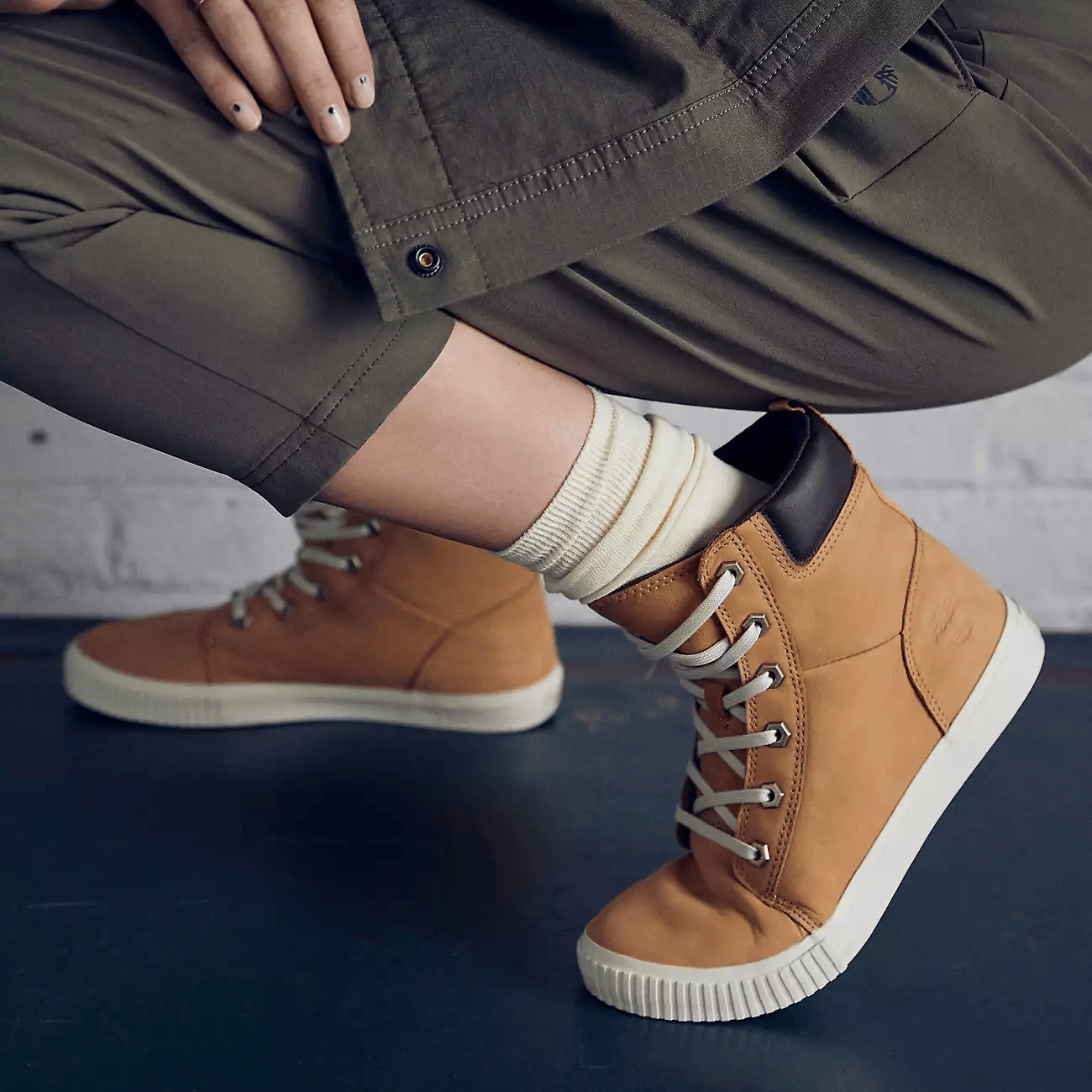A person is wearing brown high-top sneakers with cream laces and a cream-colored sole, paired with green cuffed trousers and cream socks.