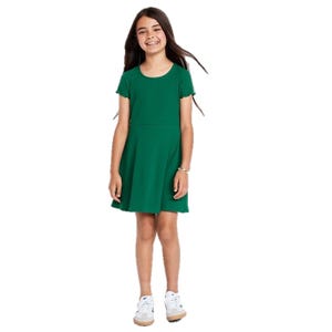 A young girl is wearing a short-sleeved green dress paired with white sneakers. She stands smiling with her hands at her sides.
