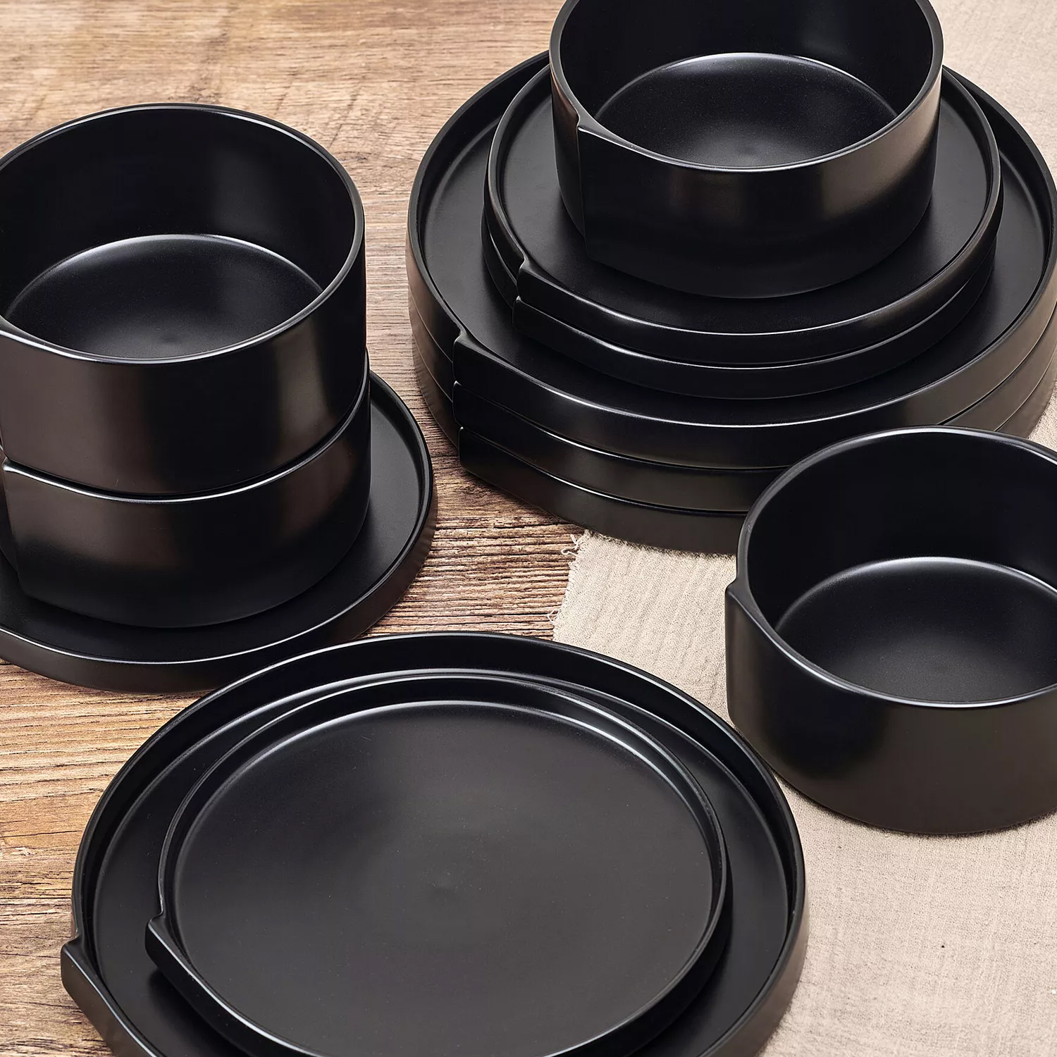 A stackable set of black dinnerware including plates, bowls, and possibly saucers is arranged on a wooden surface with a beige cloth underneath.