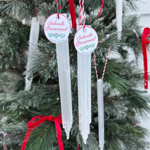Selenite ornaments hang on a Christmas tree, each labeled with a tag reading \“Selenite Ornament\“ and \“healing & cleansing,\“ featuring red and white string.