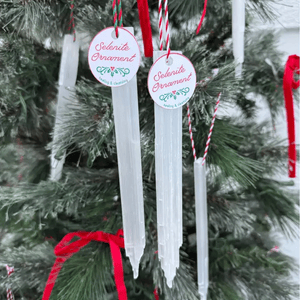Selenite ornaments hang on a Christmas tree, each labeled with a tag reading \“Selenite Ornament\“ and \“healing & cleansing,\“ featuring red and white string.