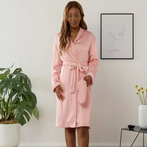 A person is wearing a pink bathrobe with a tie waist and holding a matching eye mask.