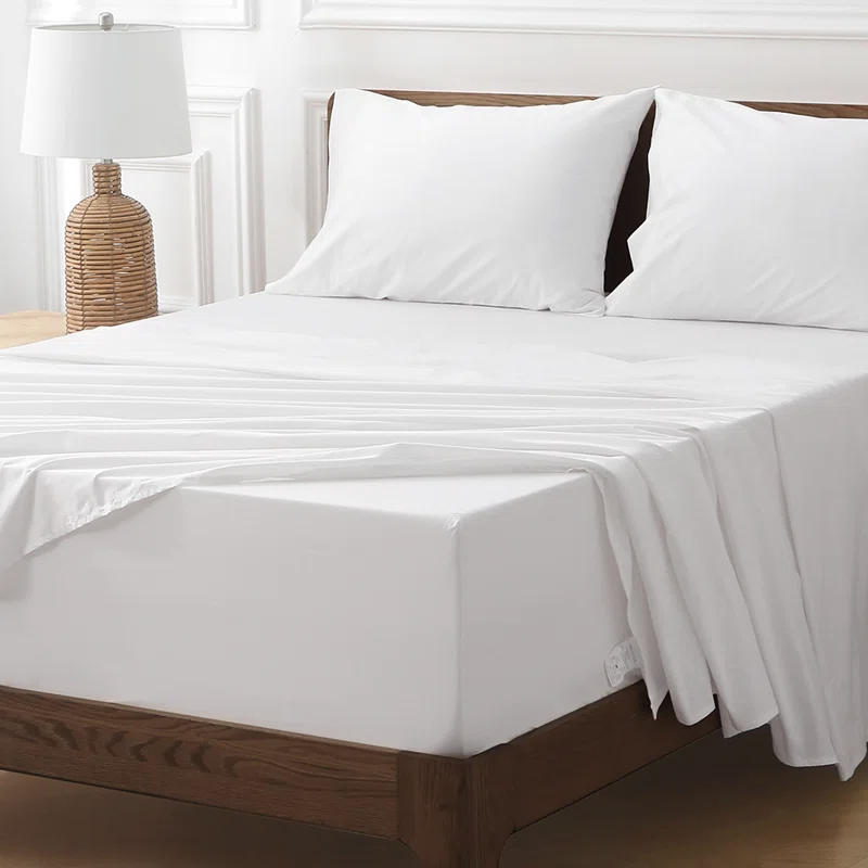 A neatly made bed with white bedding, including two pillows and sheets, on a wooden frame. A wicker-style lamp is on the bedside table.