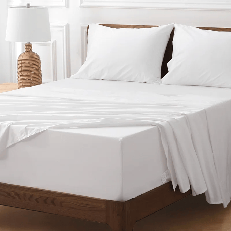 A neatly made bed with white bedding, including two pillows and sheets, on a wooden frame. A wicker-style lamp is on the bedside table.