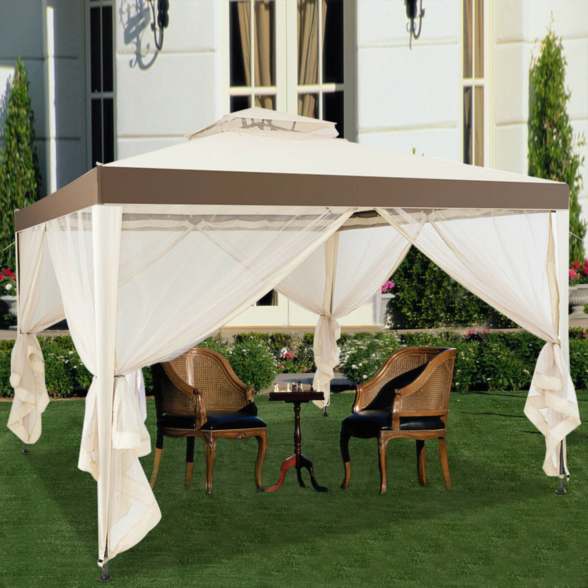 A beige and brown gazebo with sheer curtains over a small table and two wicker chairs on a lawn, set against a backdrop of a building and shrubs.