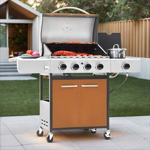 A silver and brown outdoor gas grill with five burners, a side burner with a pot, and food on the grill. It has wheels for mobility and is set on a patio area.