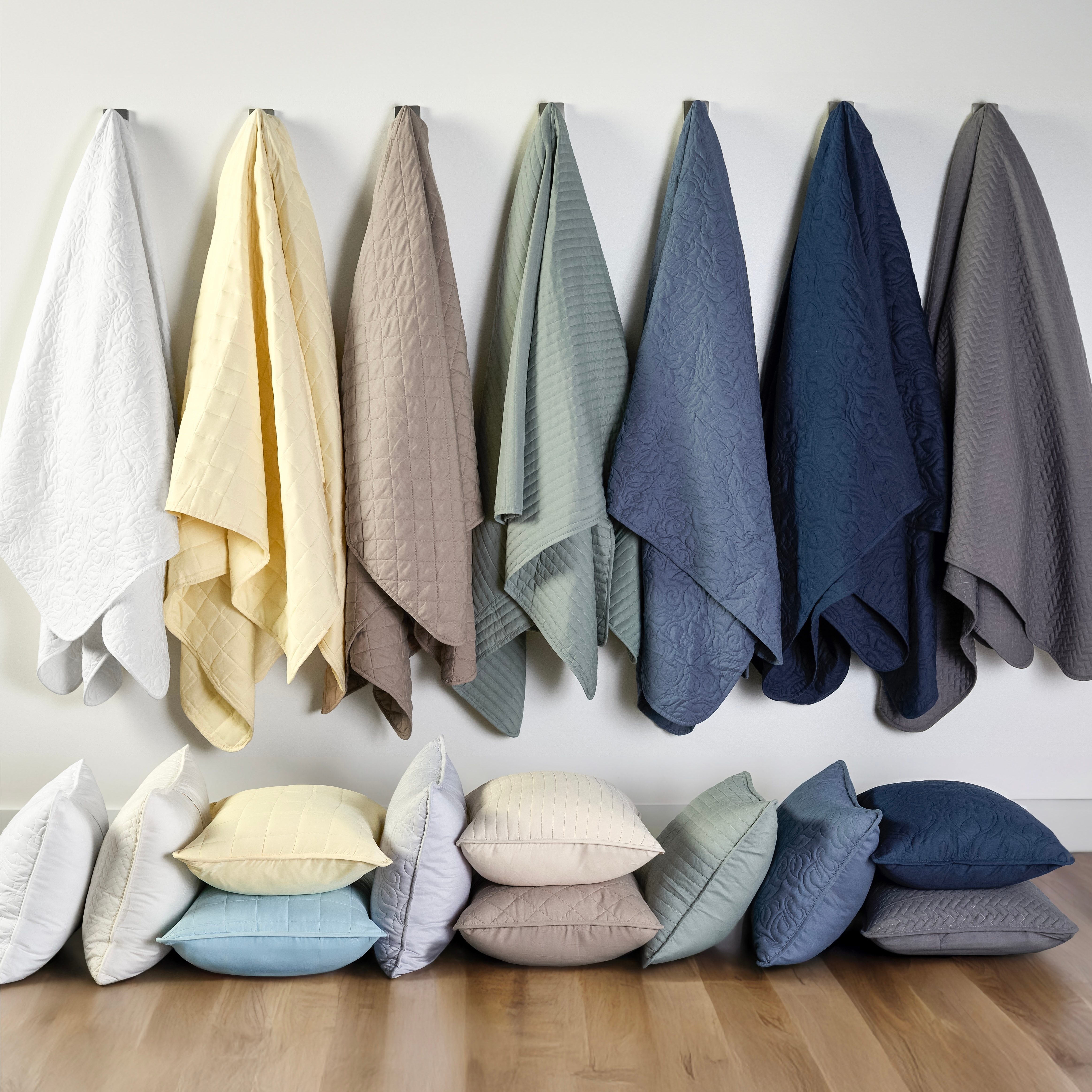 Various colored blankets hang above a row of matching pillows on a wooden floor. Colors include white, yellow, beige, green, blue, and gray, with quilts featuring subtle patterns.
