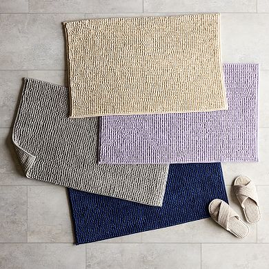 Four chenille bath rugs in different colors are overlapped on a tiled floor, accompanied by a pair of slippers at the side.