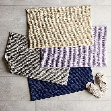 Four chenille bath rugs in different colors are overlapped on a tiled floor, accompanied by a pair of slippers at the side.