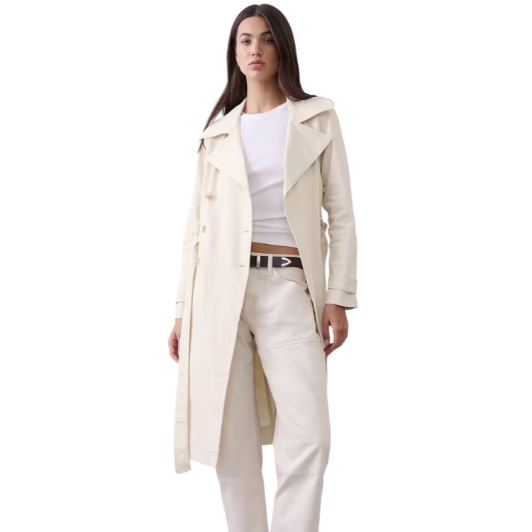 A woman wears a cream-colored trench coat over a white top and cream pants, paired with a black belt and brown ankle boots.