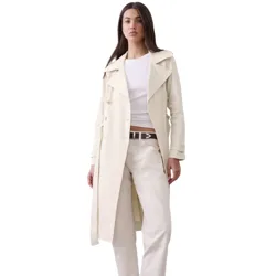 A woman wears a cream-colored trench coat over a white top and cream pants, paired with a black belt and brown ankle boots.