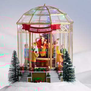 A festive figurine displays a holiday scene with musicians in Santa hats playing instruments inside a decorative gazebo. \“Merry Christmas\“ is written on a banner, surrounded by steps, trees, and a snowy landscape, creating a warm holiday ambiance.