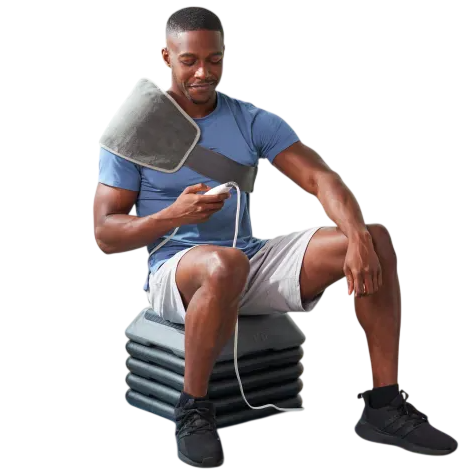 A man uses an adjustable heating pad wrapped around his shoulder while sitting on a stack of exercise step platforms. Nearby are a foam roller and a striped resistance band on a yoga mat.
