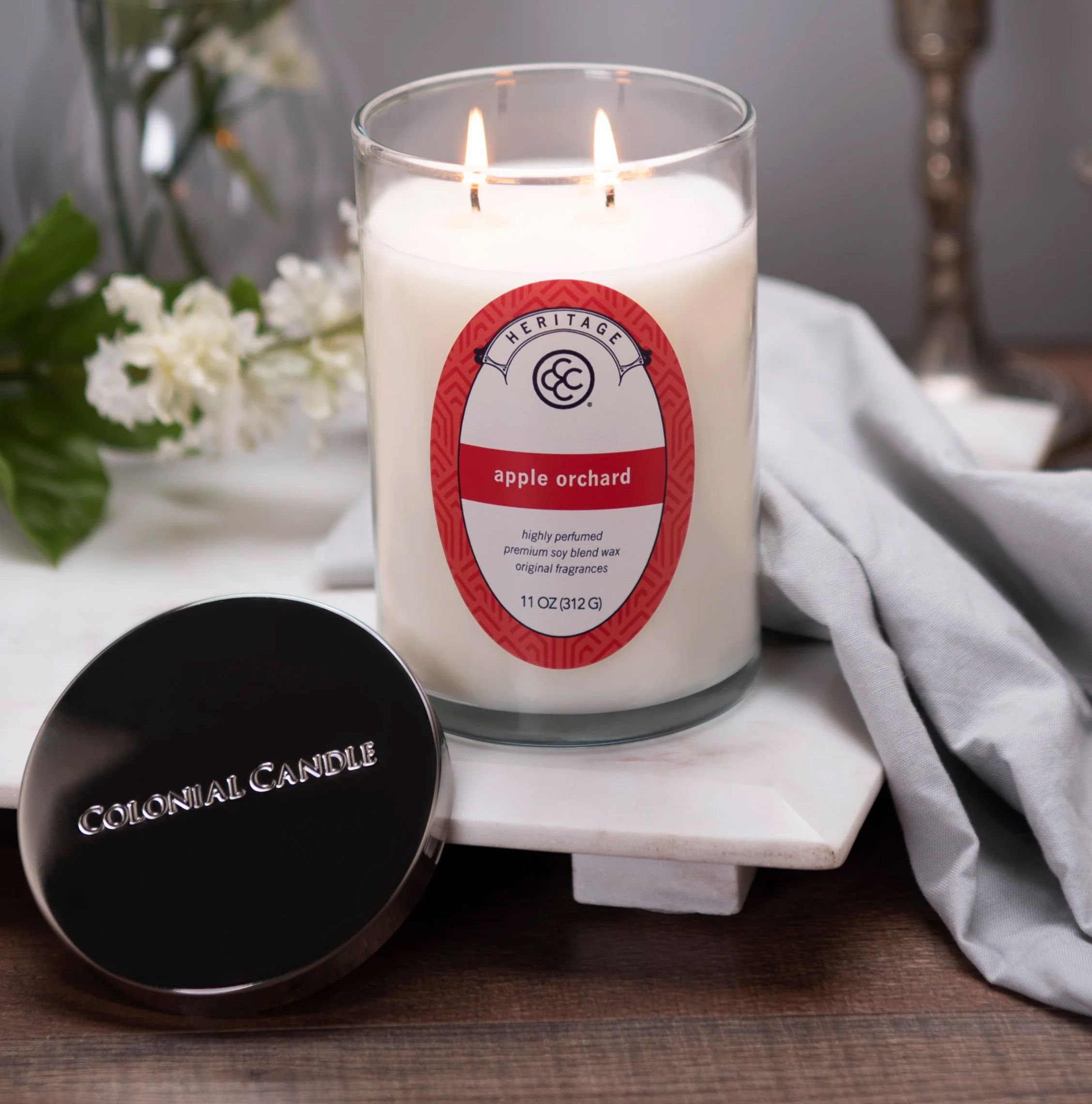 A Colonial Candle \“Apple Orchard\“ scented candle with two wicks, featuring a highly perfumed premium soy blend wax in an 11 oz (312g) glass container. The lid is placed beside the candle.