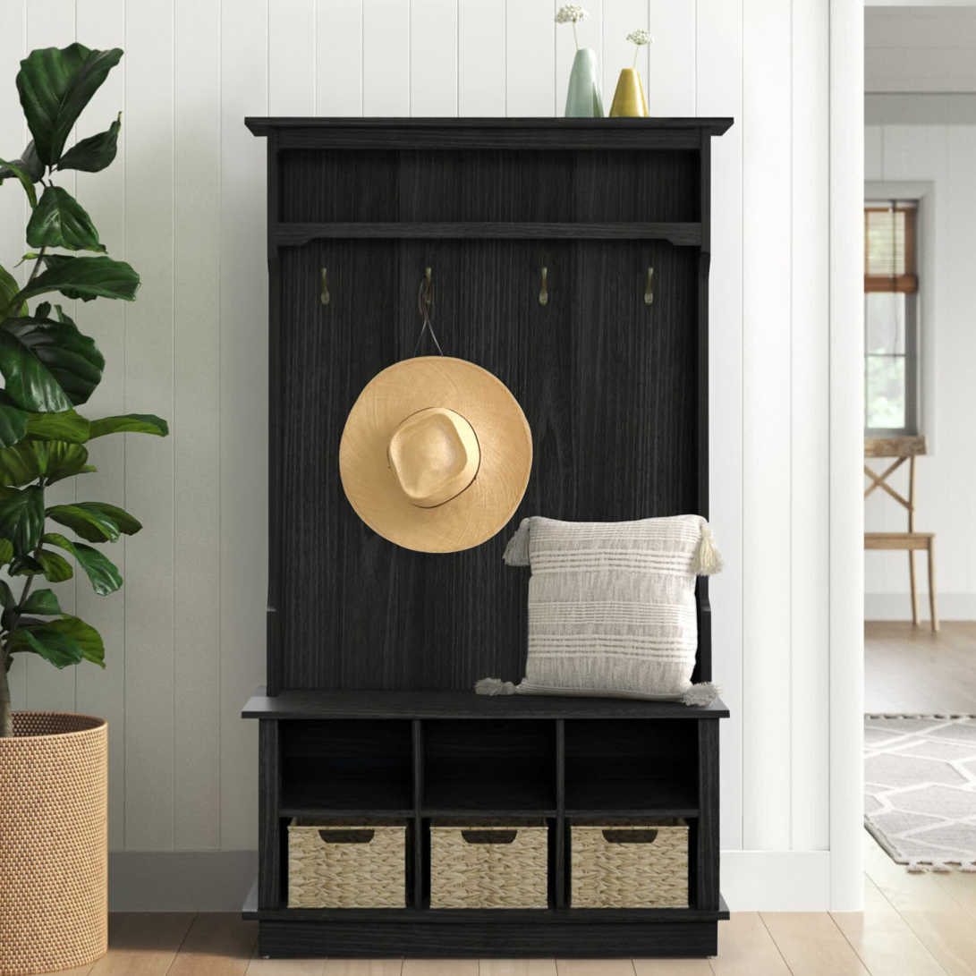 Black hall tree with three hooks, a cushioned bench, and three wicker baskets for storage. A straw hat hangs on one hook, and a textured pillow is on the bench. A large potted plant is nearby, and two decorative vases sit on top.