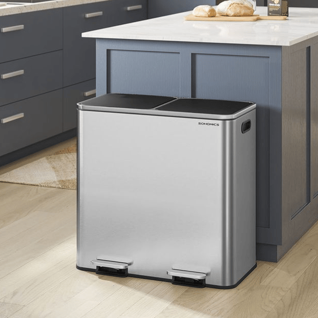 A modern kitchen features a dual-compartment, stainless steel trash can with foot pedals, alongside a blue and white cabinetry setup, a countertop with bread and a plant, and an oven.