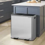 A modern kitchen features a dual-compartment, stainless steel trash can with foot pedals, alongside a blue and white cabinetry setup, a countertop with bread and a plant, and an oven.