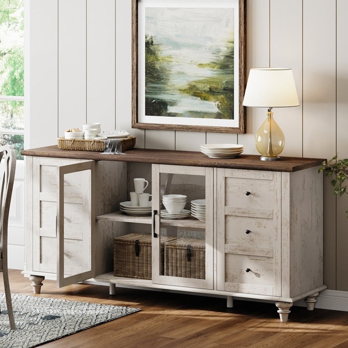 A farmhouse-style sideboard features a dark wood top and distressed white finish, with multiple drawers and glass door cabinets, displaying dishes and baskets. A table lamp and decorative items are placed on top, beneath a scenic painting.