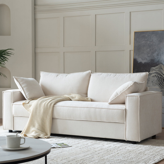 Beige sofa with two large back cushions and two side pillows, draped with a knitted throw. A round coffee table with a cup and a magazine on the floor are in the foreground, and a framed artwork leans against a paneled wall.