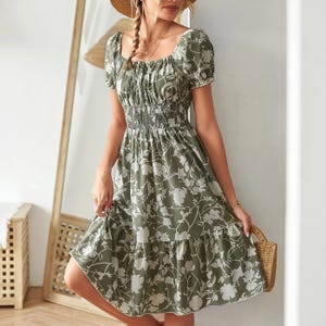 A woman wearing a green and white floral dress with puffed short sleeves, a smocked waist, and a tiered skirt. She accessorizes with a straw hat and holds a woven bag.