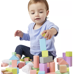 A toddler plays with colorful wooden building blocks, including various shapes like cubes, cylinders, and triangular prisms, encouraging creative play and motor skills development.