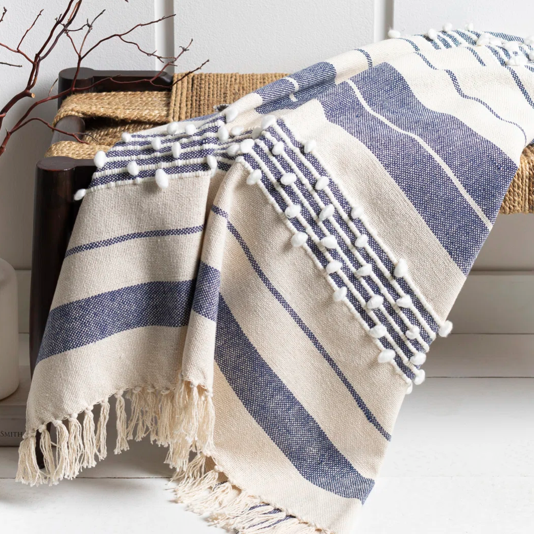 A blue and white striped, fringed throw blanket with pom-pom detailing is draped over a bench next to a vase with decorative twigs.