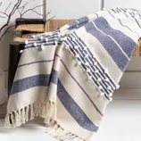 A blue and white striped, fringed throw blanket with pom-pom detailing is draped over a bench next to a vase with decorative twigs.