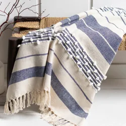 A blue and white striped, fringed throw blanket with pom-pom detailing is draped over a bench next to a vase with decorative twigs.