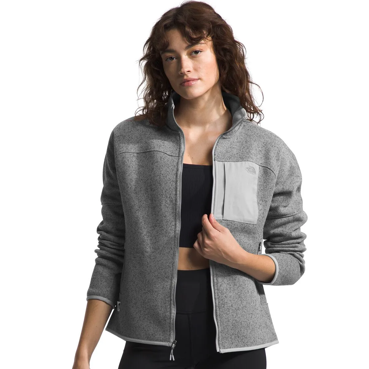 A woman wearing a grey zip-up jacket with a light grey pocket and black sports bra.