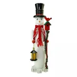 A snowman figurine with a black top hat, red scarf, and two beaded garlands holds a lantern in one hand and a stick with a red birdhouse and cardinal in the other.