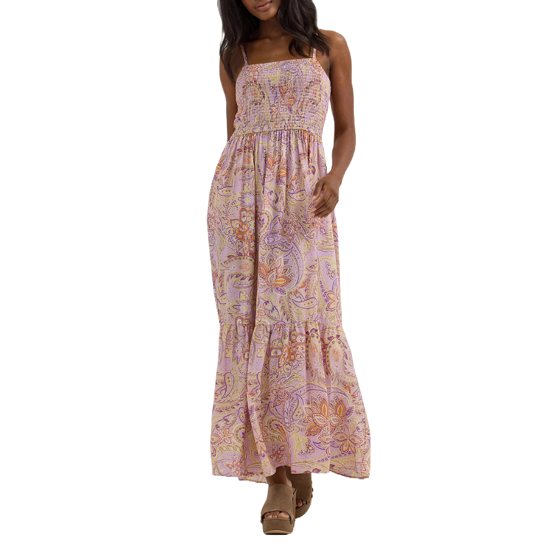 A woman is wearing a sleeveless, ankle-length dress with a smocked top and paisley print in shades of pink, orange, and purple, paired with tan platform sandals.