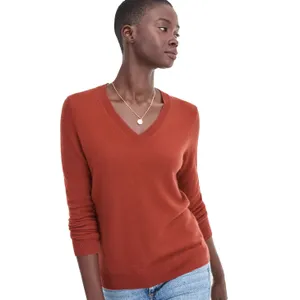 A person wearing a rust-colored V-neck sweater and light blue jeans, accessorized with a simple gold necklace.