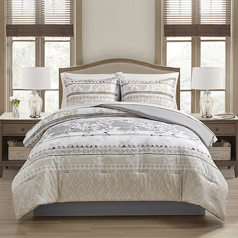 A neatly made bed features a patterned comforter and two matching pillows in beige and gray tones. The setup includes a wooden headboard, a pair of glass-base lamps, and wooden nightstands with woven baskets beneath.